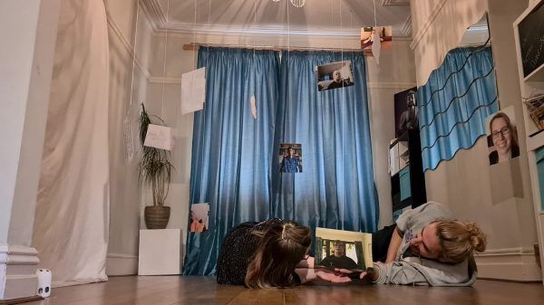 An image of the artists practicing their work in progress. Jen and David lie on the floor, the camera is at their heads and above photos of their friends dangle from the ceiling.