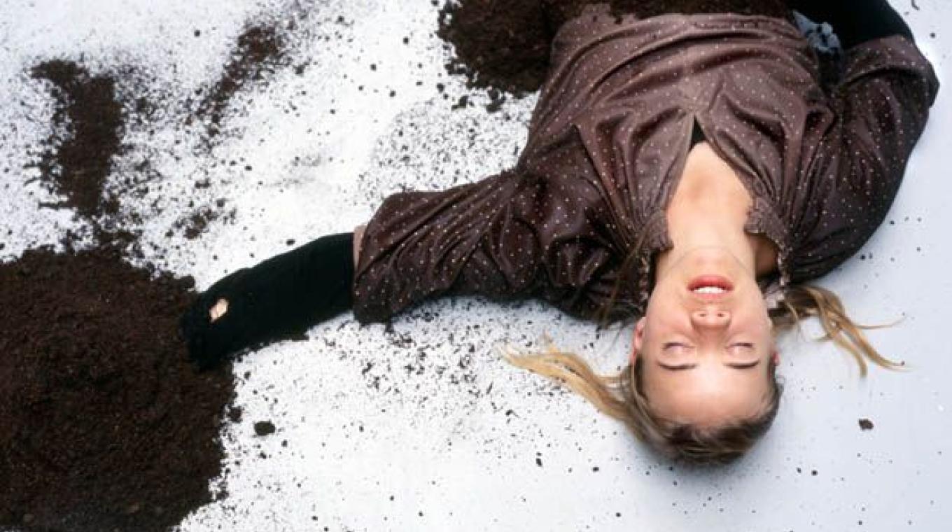 A woman lying on a white floor with her eyes shut and mounds of soil over her hands