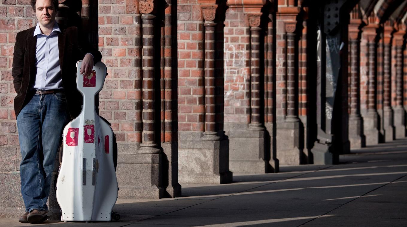 A man stood under a cloister with his hand propped on the top of a cello case