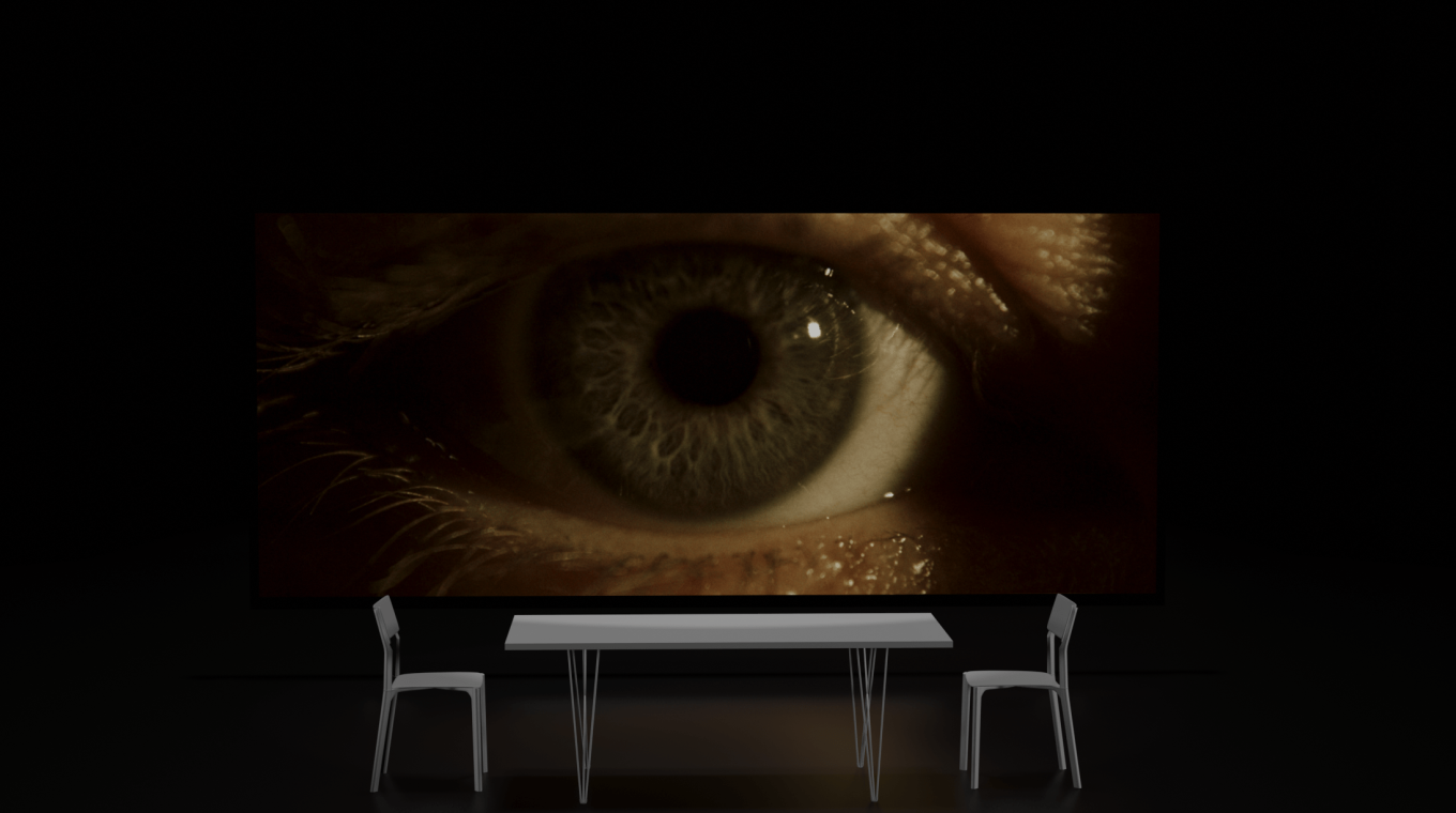 A white table with two chairs (one either side) are placed in front of a screen which is showing an extreme close up of an eyeball and eyelid.