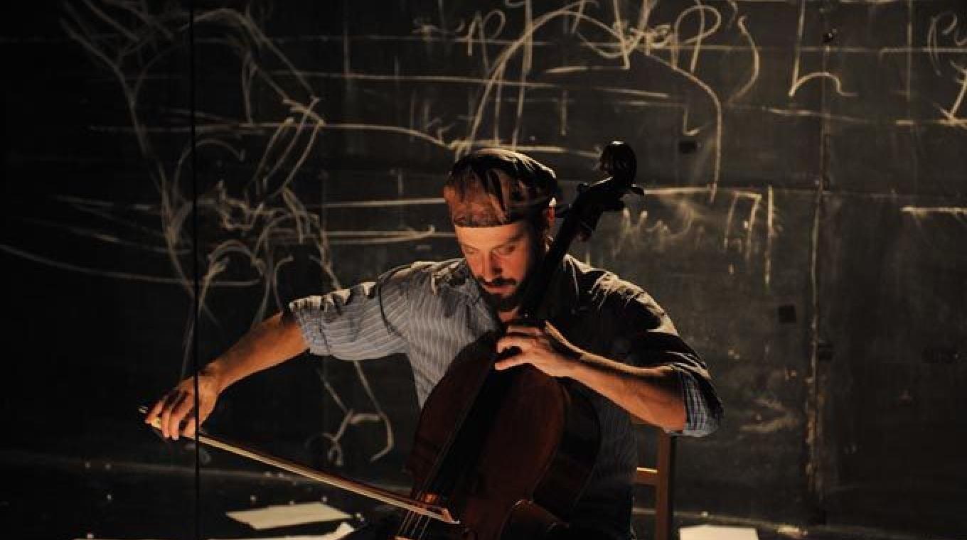A man playing the cello under a naked light bulb with sheets of paper on the floor