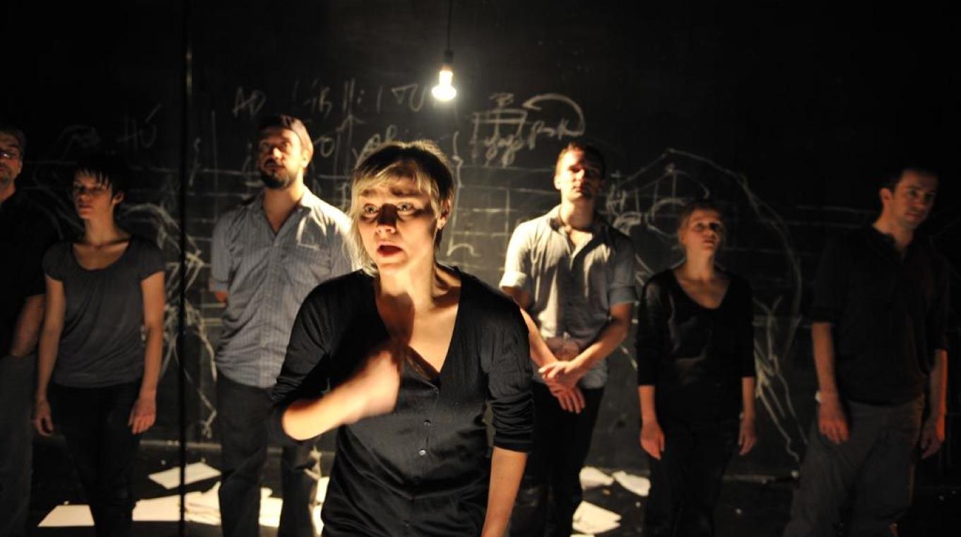 A stage photo of the company cast with an actress at the front under a naked light bulb.