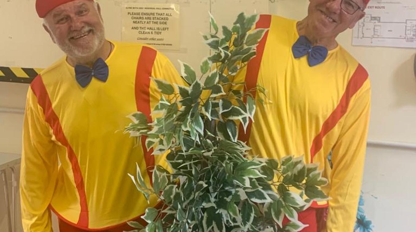 Two white males wearing a yellow and red costume leaning out either side of a plant.