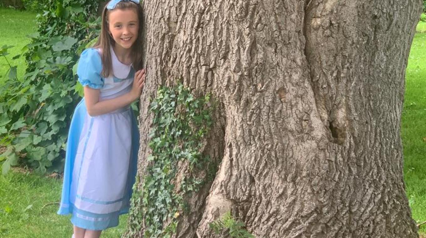 A young girl dressed as Alice in Wonderland stood leaning against a tree