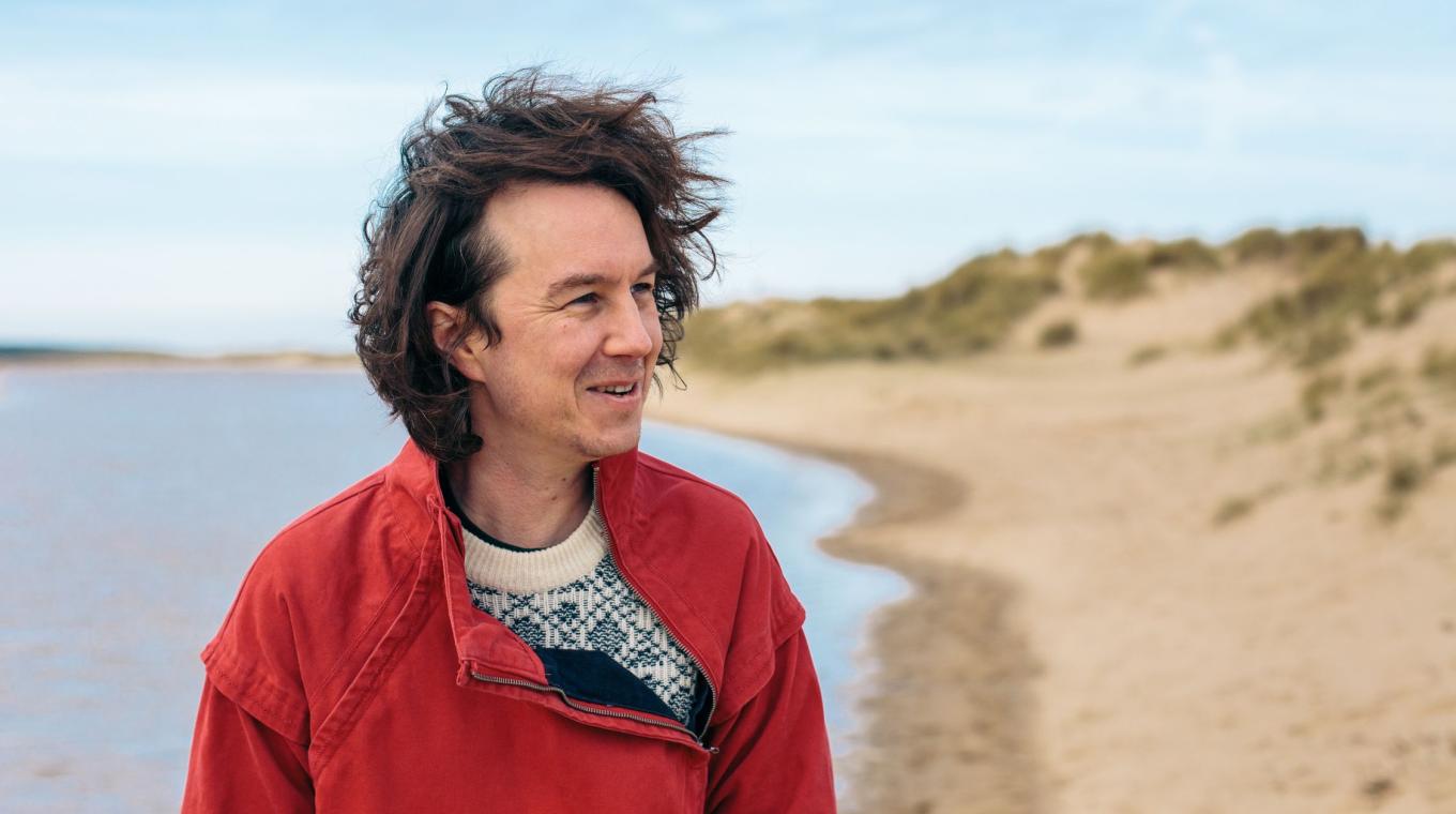 A white man with medium length hair wearing a red jacket stood on a beach looking to the right of frame