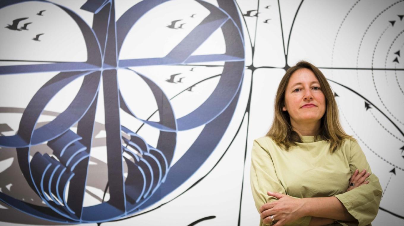 A white woman with shoulder length hair standing to the right with her arms folded looking at the camera. With artwork behind her featuring birds and circles