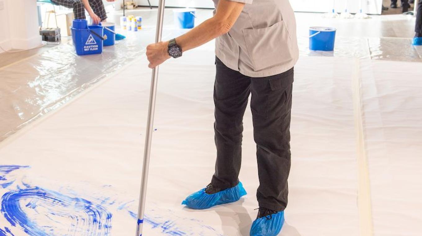 A female cleaner standing on canvas using a cleaning mop to paint blue marks on the white canvas