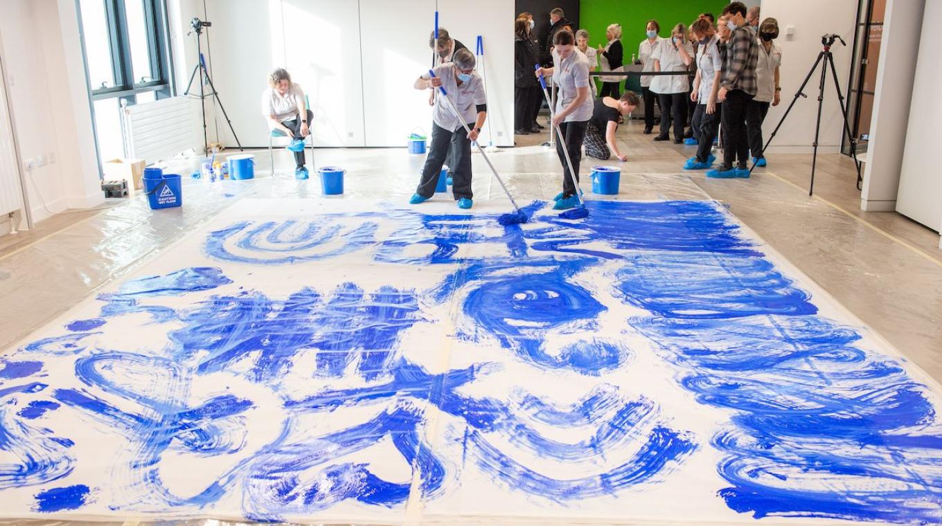 Two female cleaners standing on canvas using a cleaning mop to paint blue marks on the white canvas