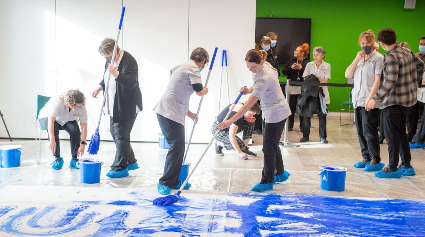 Two female cleaners standing on canvas using a cleaning mop to paint blue marks on the white canvas