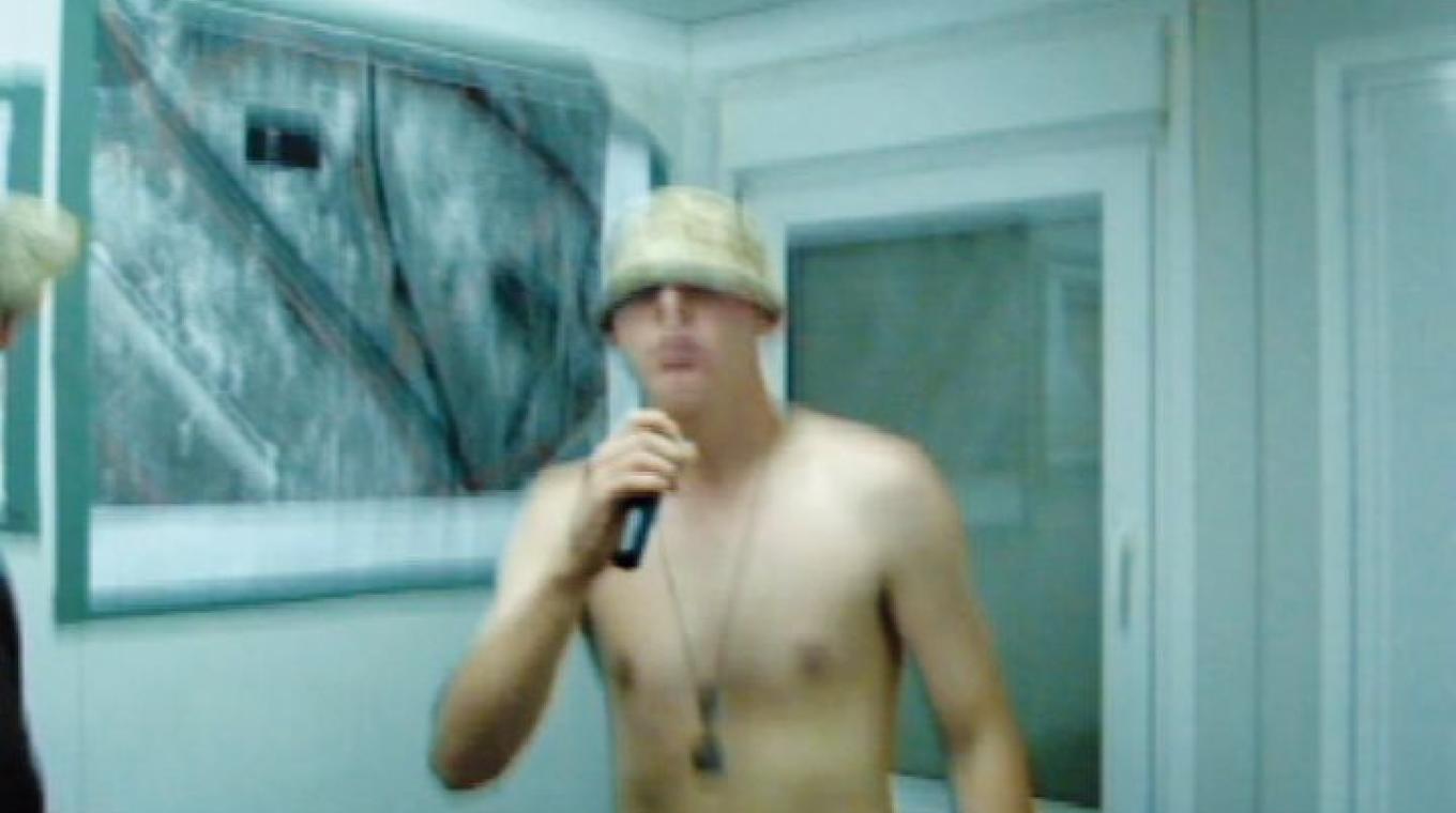Blurry Photo of a bare chested man with a hat and a 'dog tag' around his neck - holding a microphone