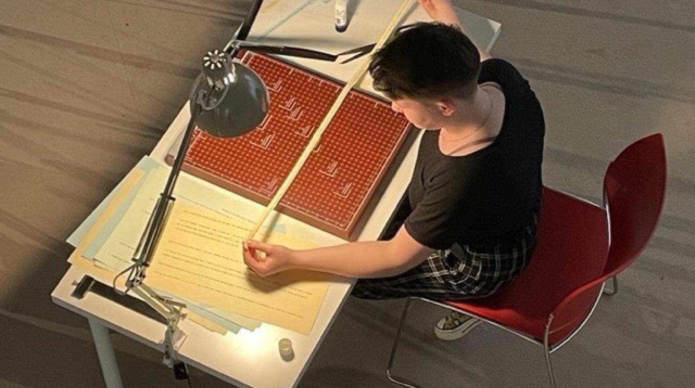 A photo from above showing a young woman sat on a red chair in front of a desk holding out a long thin strip of paper