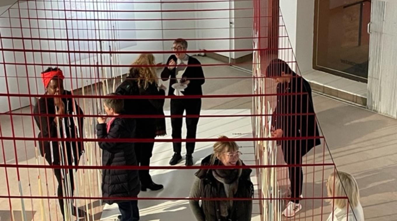 A photo from above of people standing in a gallery chatting with thin strips of paper hanging along an open tunnel