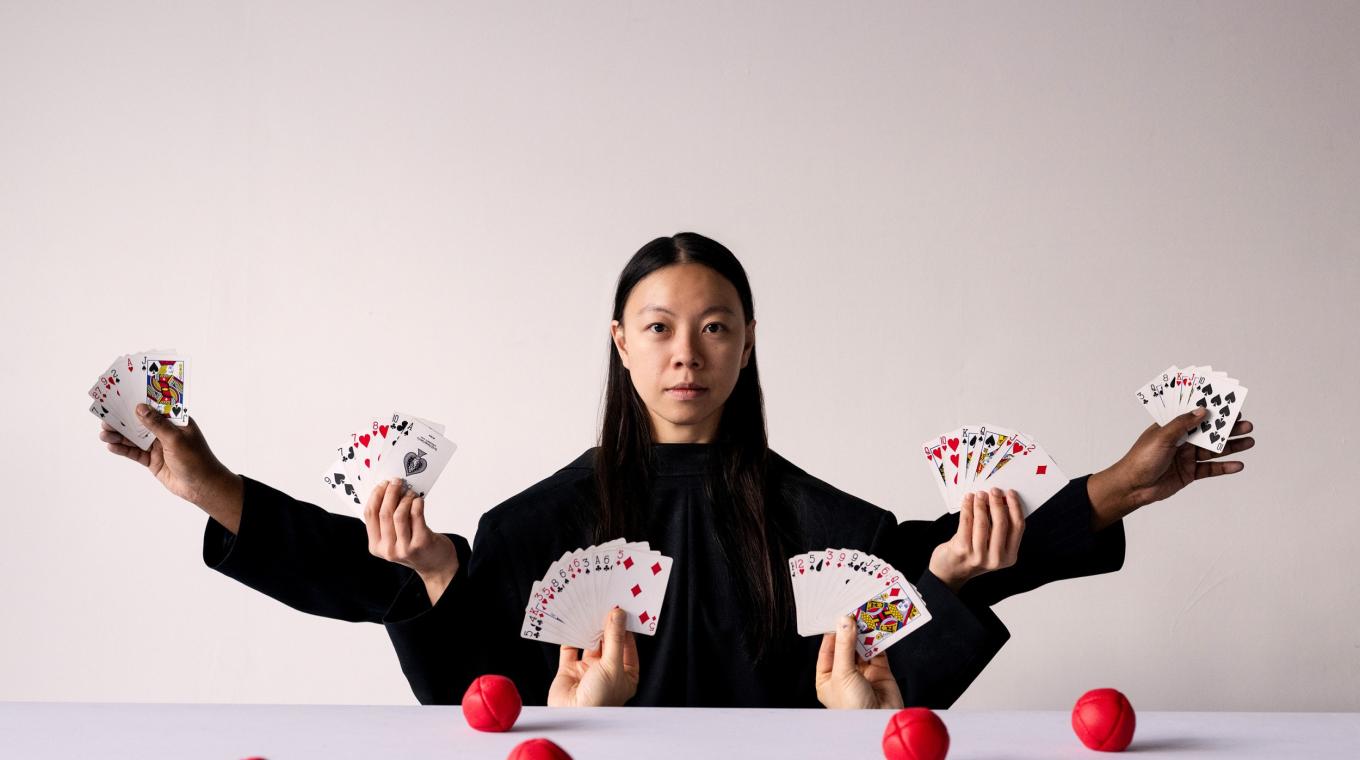 A woman with her arms stretched out holding playing cards in each hand. Four more hands reach up from below a table also holding hands