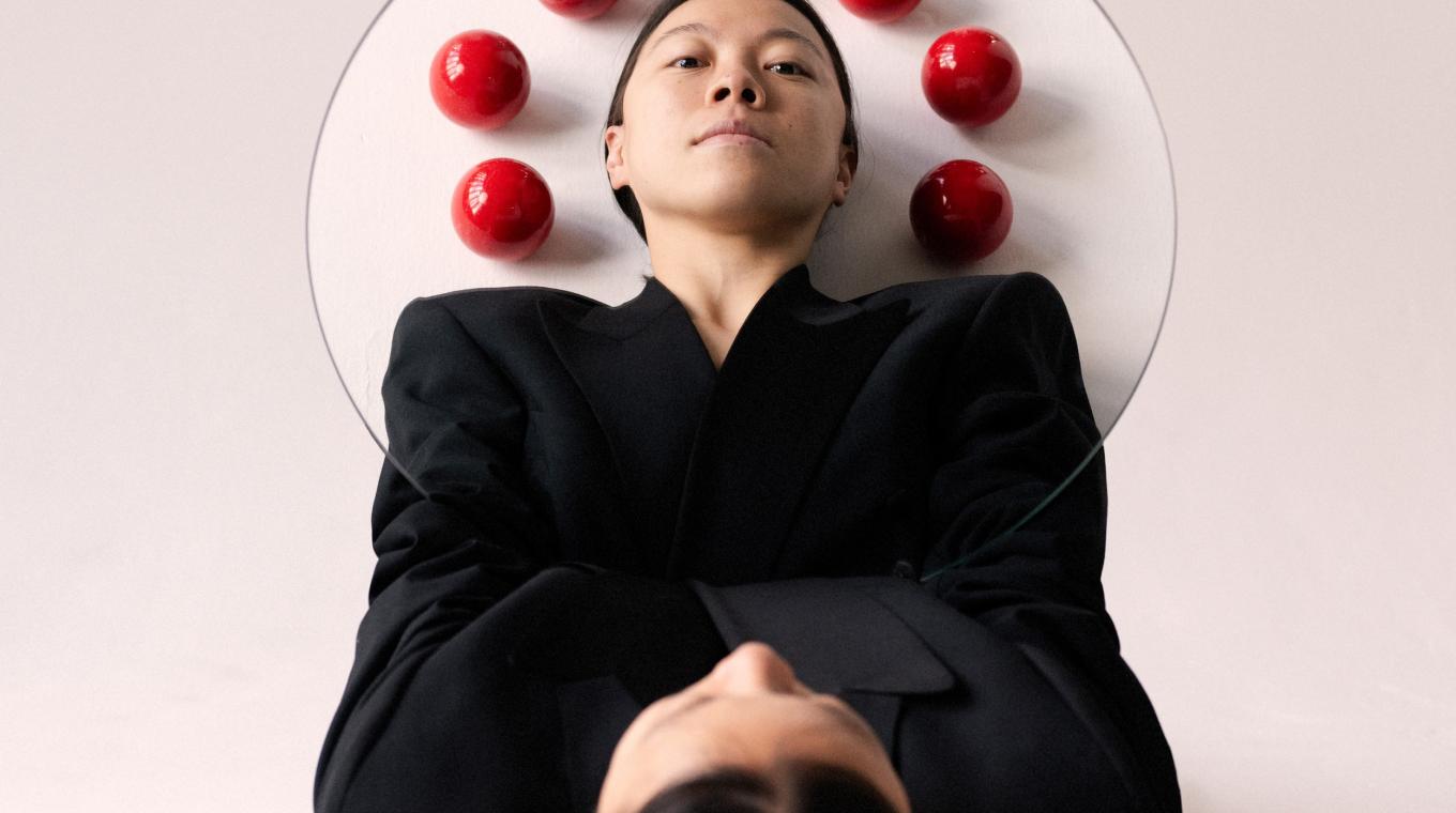 A woman lying down with a circular mirror refelcting her face and seven juggling balls