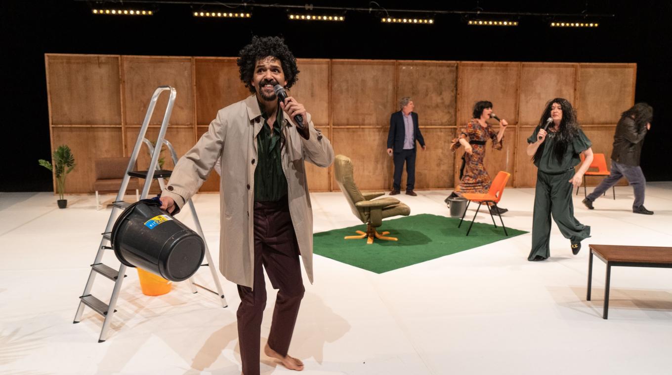 A man in a trench coat holding a bucket with people and props in the background