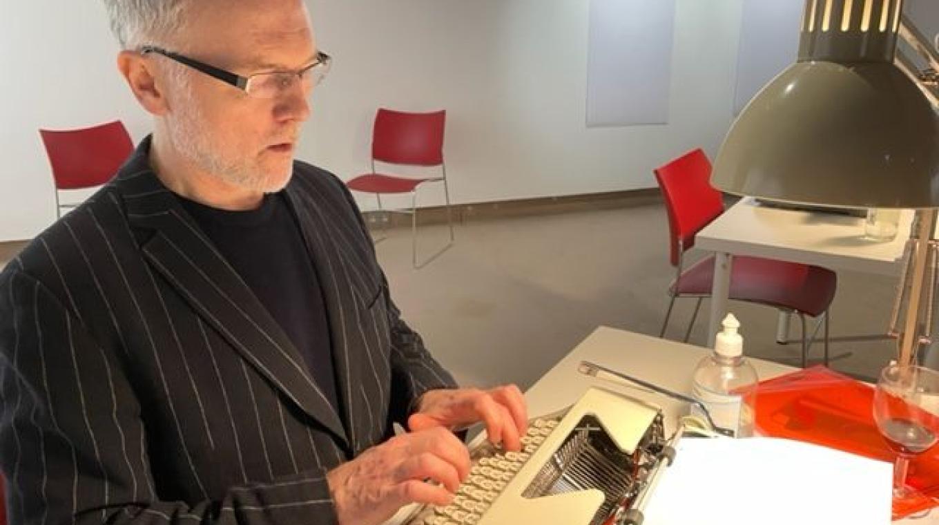A man wearing glasses with grey hair sitting at a desk typing on an old typewriter with a desk lamp above the typewriter