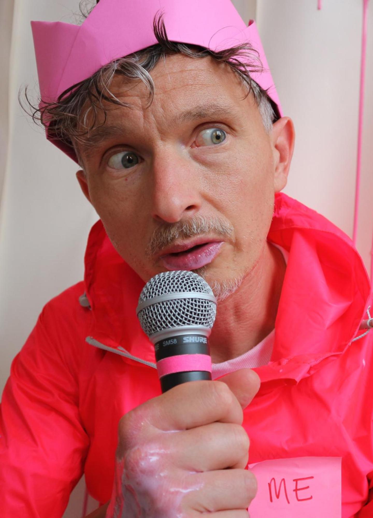 A man in a pink party hat and pink rainciat with a microphone and pink paint dripping down the wall behind him