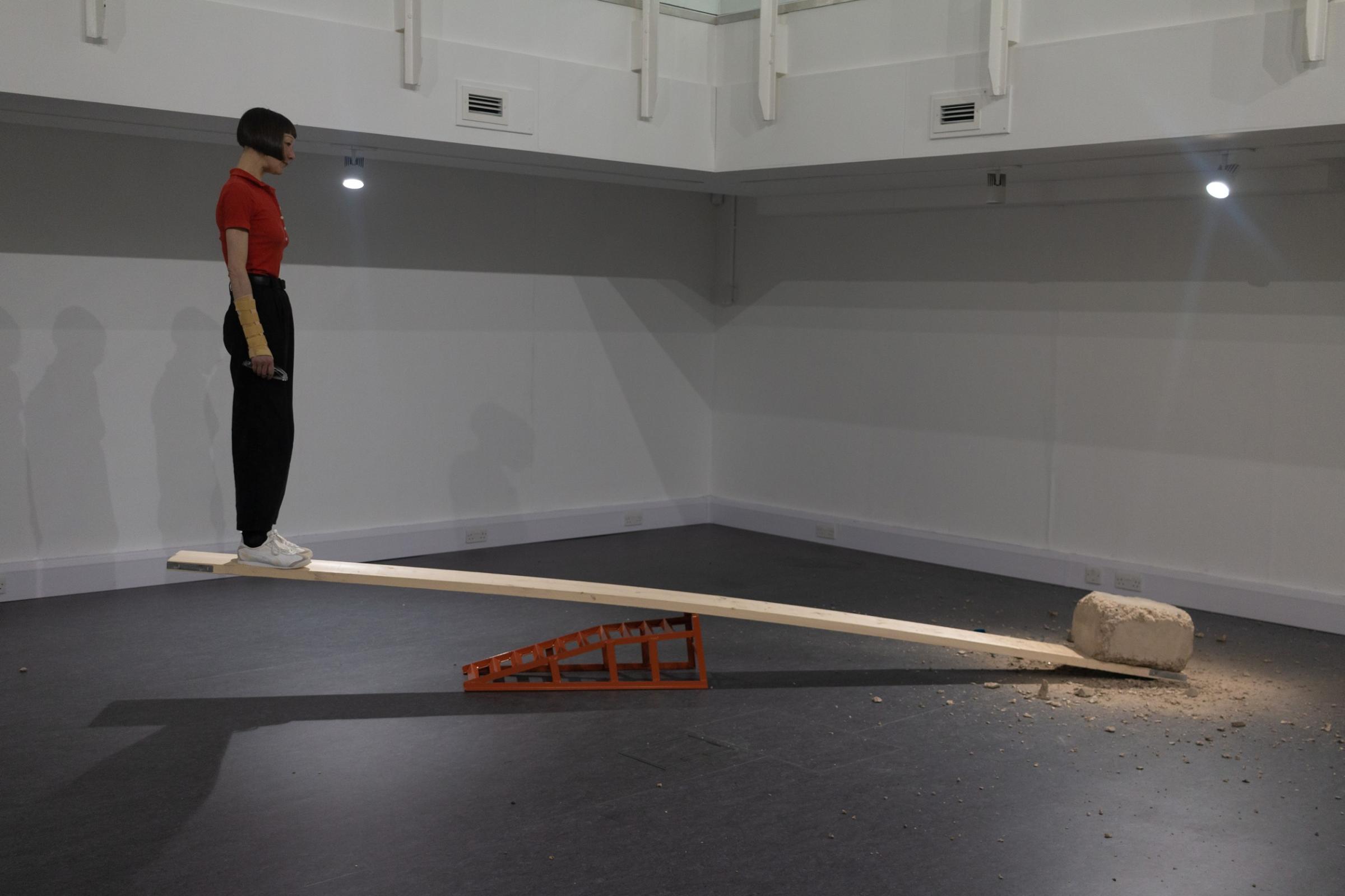 A woman standing on a plank, looking at a block of concrete at the other end of the plank that has been chipped away. The seesaw is weighted down by the concrete so the woman's end is off the ground.