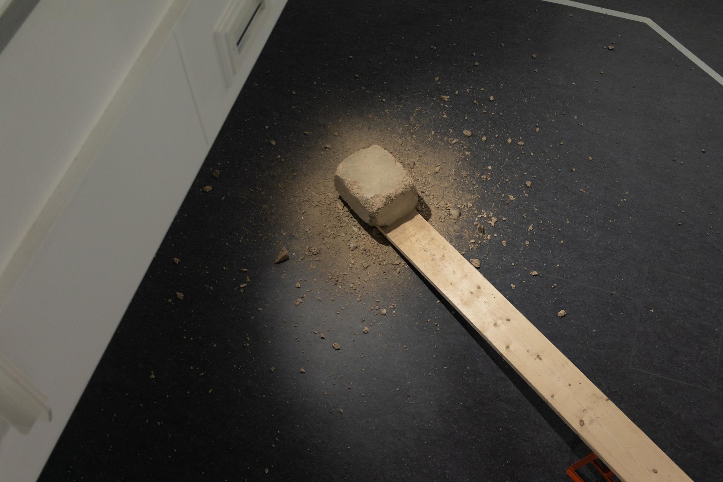 Aerial view of a block of concrete that has been chipped away with debris across the floor. The concrete block rests on the end of a plank. It is located in an art gallery.