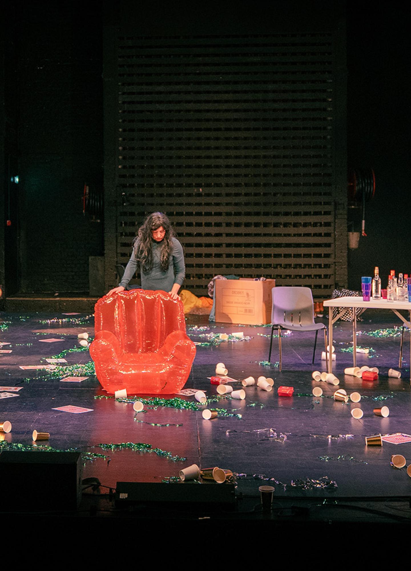 A chaotic scene on stage with three people, two inflatable chairs and a table with cups and decorations strewn across the floor