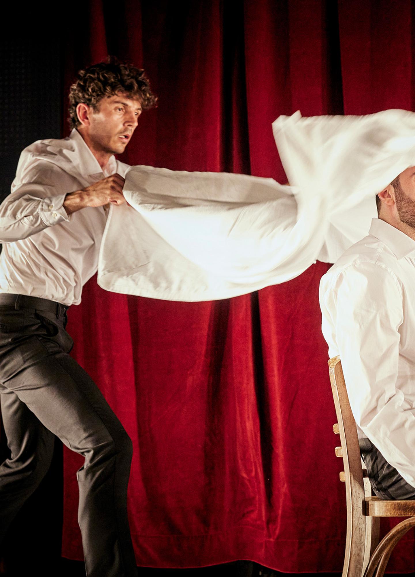 A waiter throwing a white table cloth over the head of a diner sitting at a table with a red curtain behind