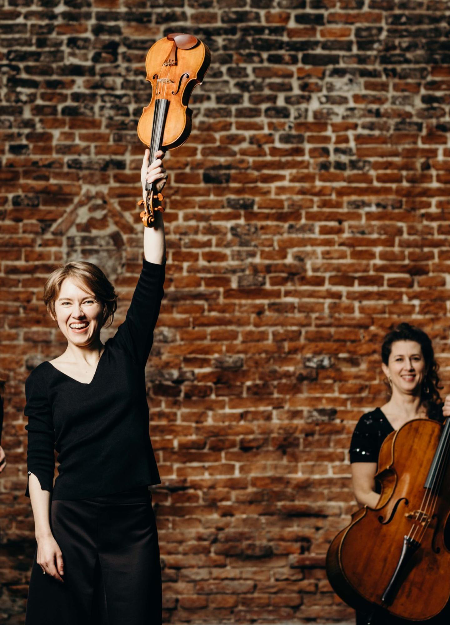 Four women with string instruments. The one at the front is raising a violin in the air.