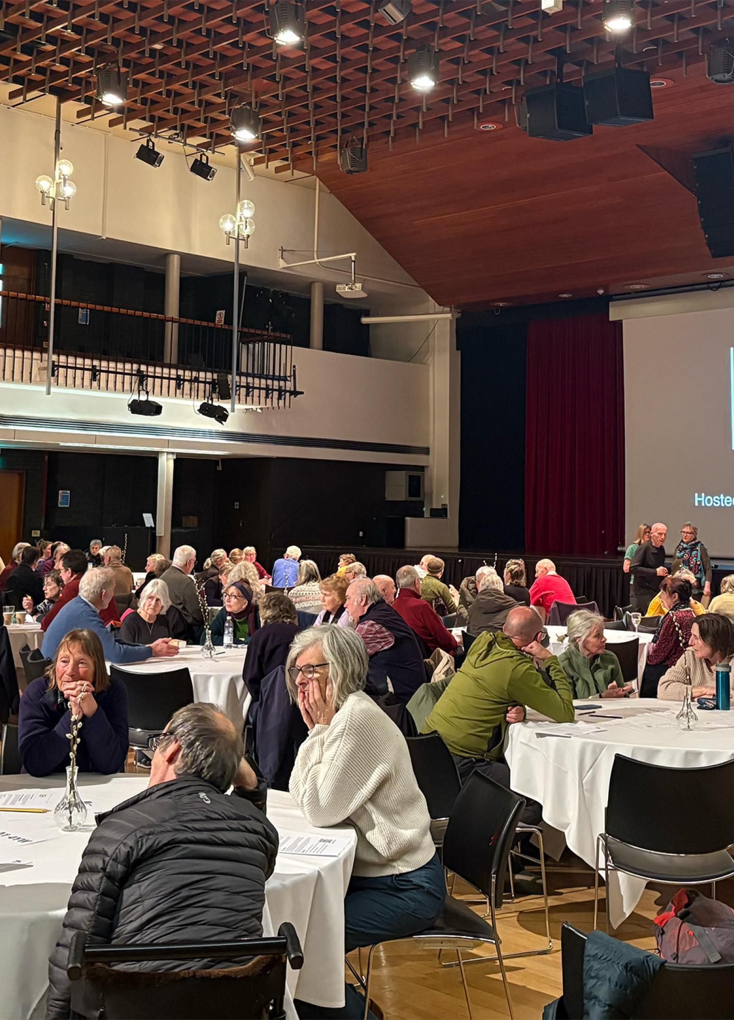 A hall full of people at round tables talking with a big screen that says "People's Emergency Briefing"