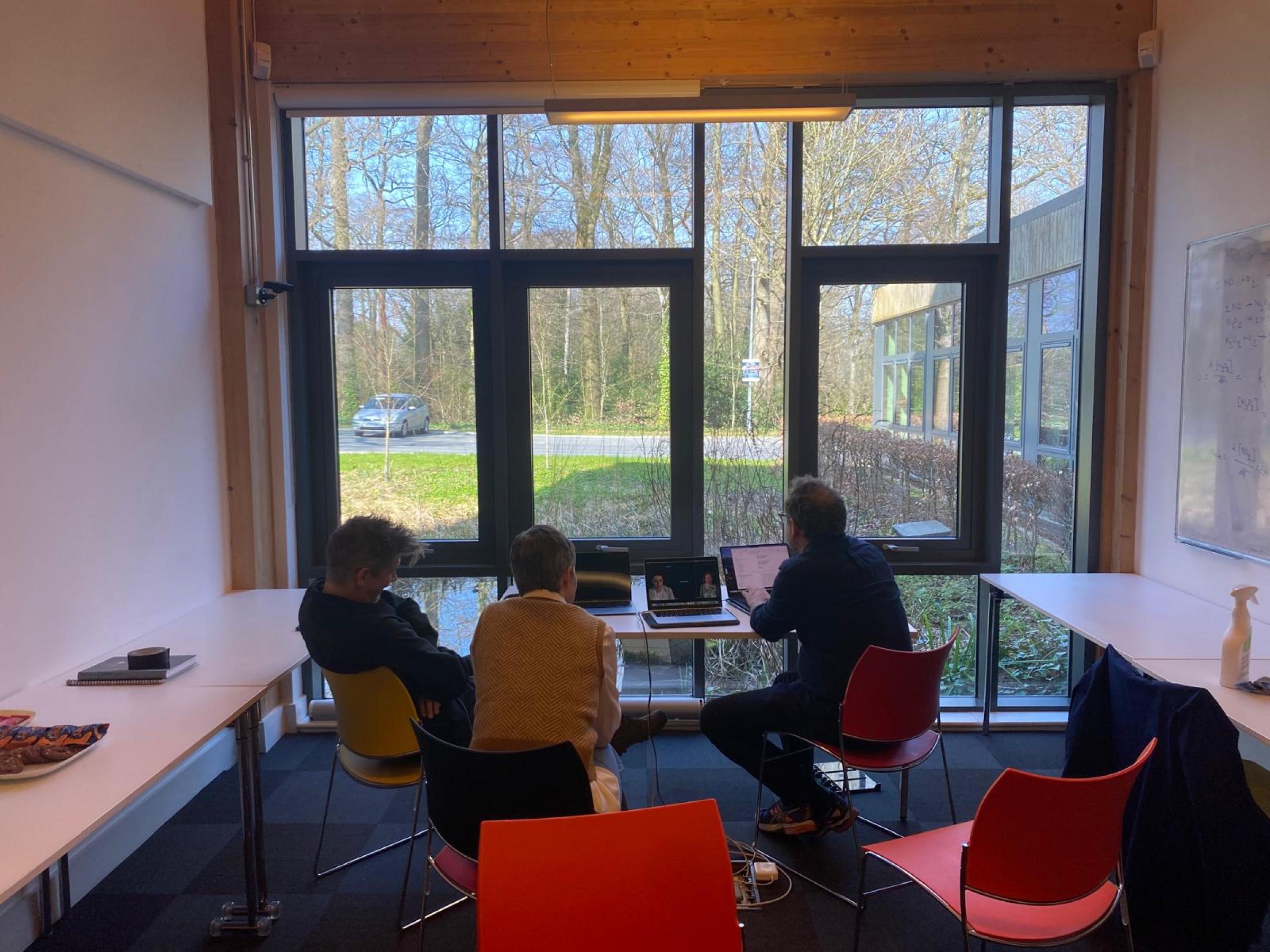 Three people sitting around a laptop in front of a big window with greenery outside