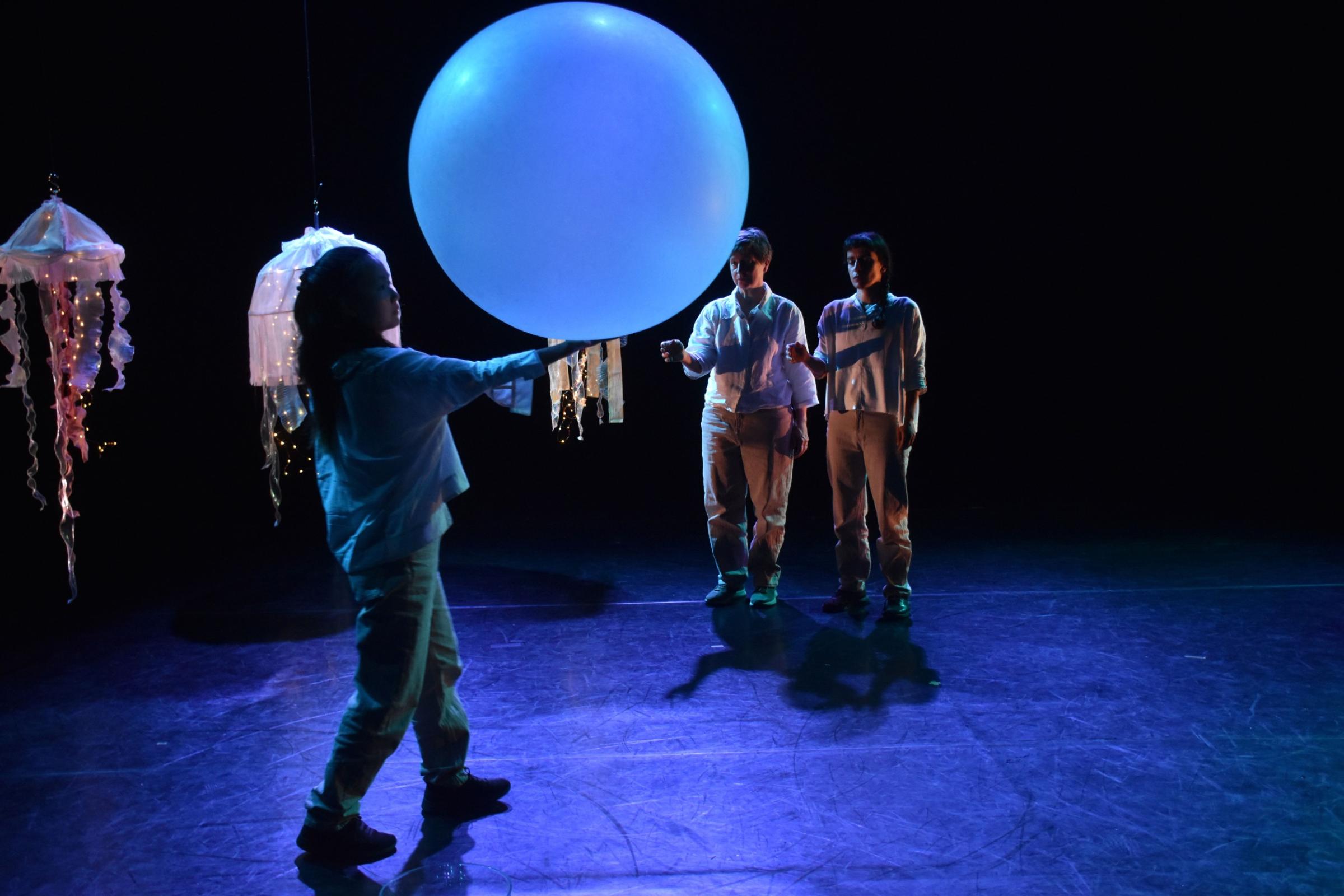 Three performers on stage one holding a large spherical baloon with ocopus textiles hanging from above