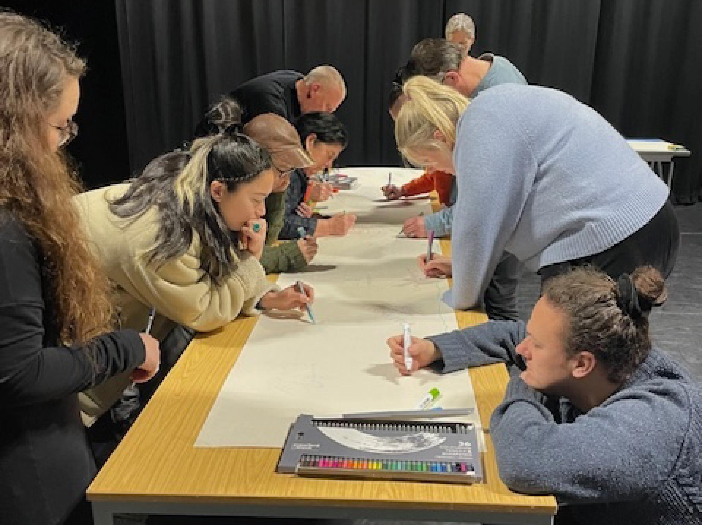 A group of people writing on a long piece of paper across a table