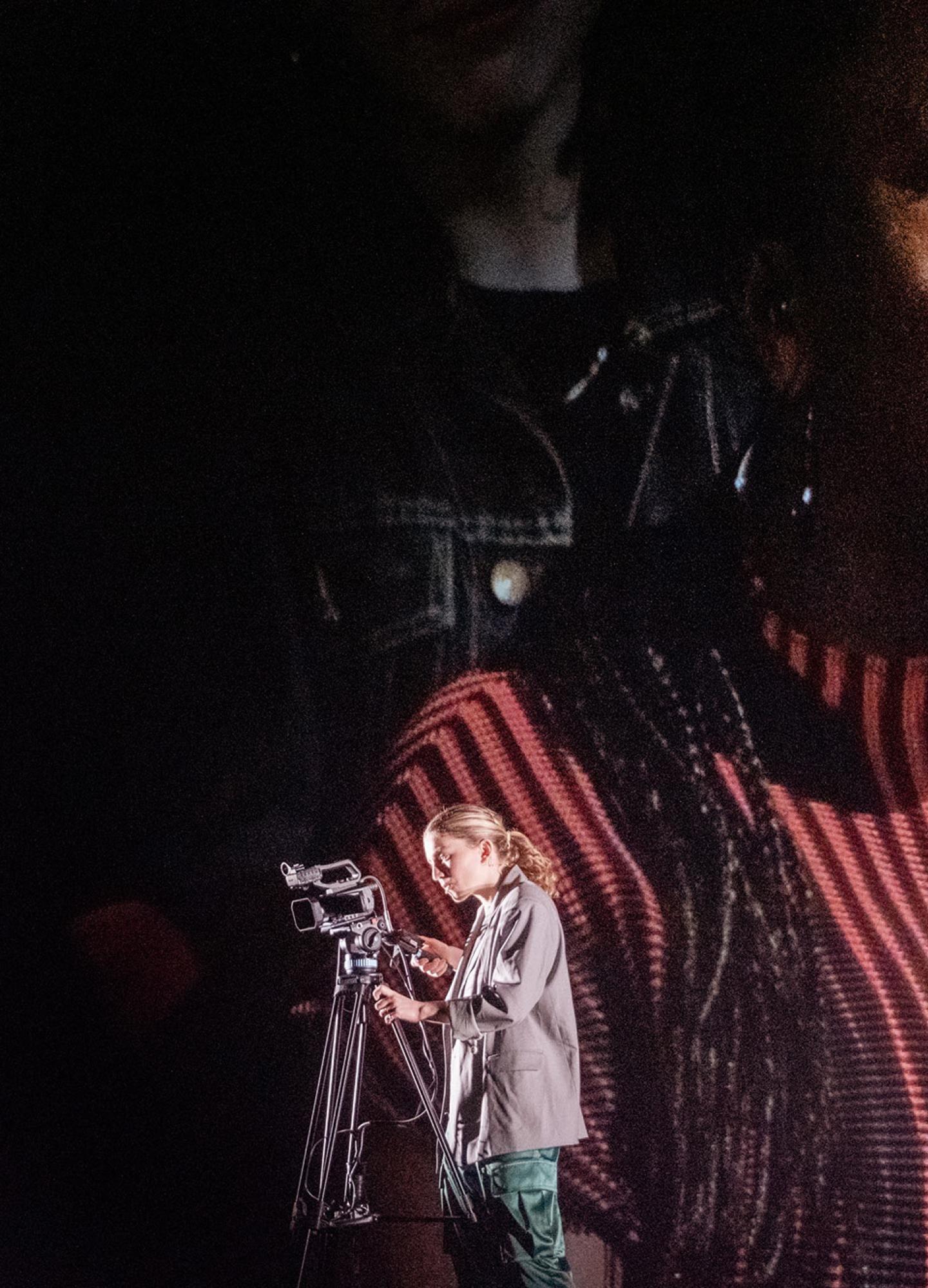 A person in a red top on a big screen smiling with a person holding a video camera in front of the screen