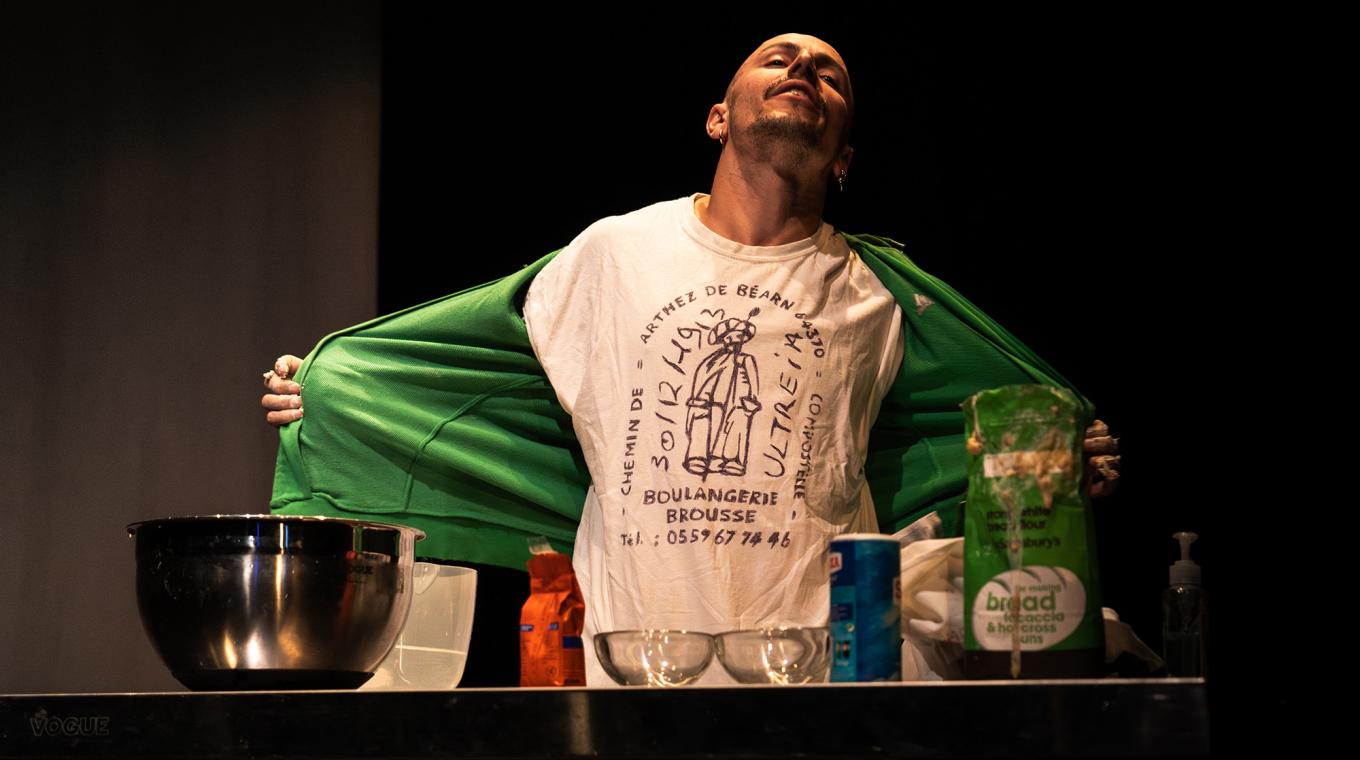 A man revealing a t-shirt featuring the words Boulangerie Brousse standing in front of a table full of baking bowls and flour