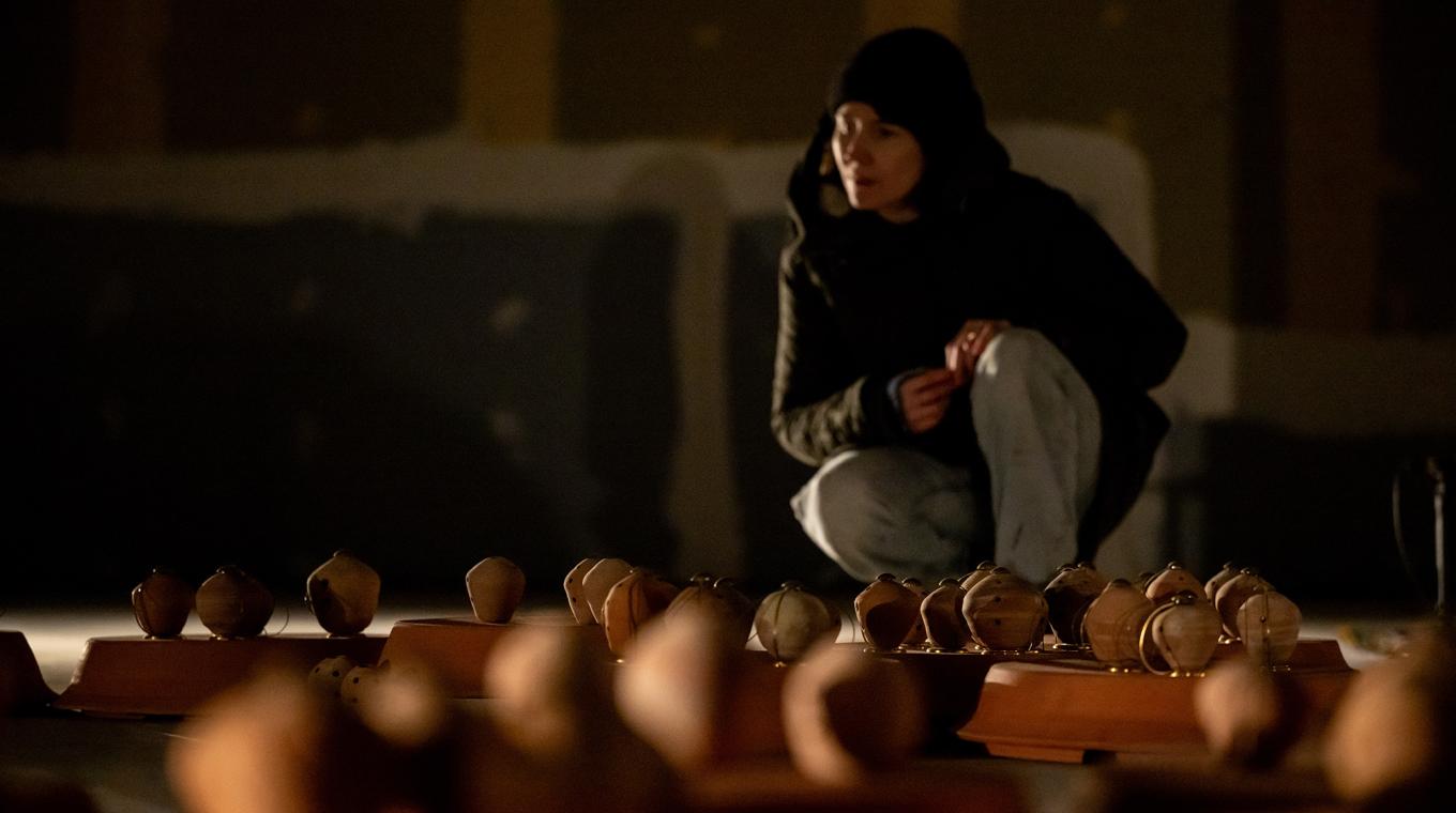 Someone with a hood up crouching down looking at around 30 small clay pots with holes in