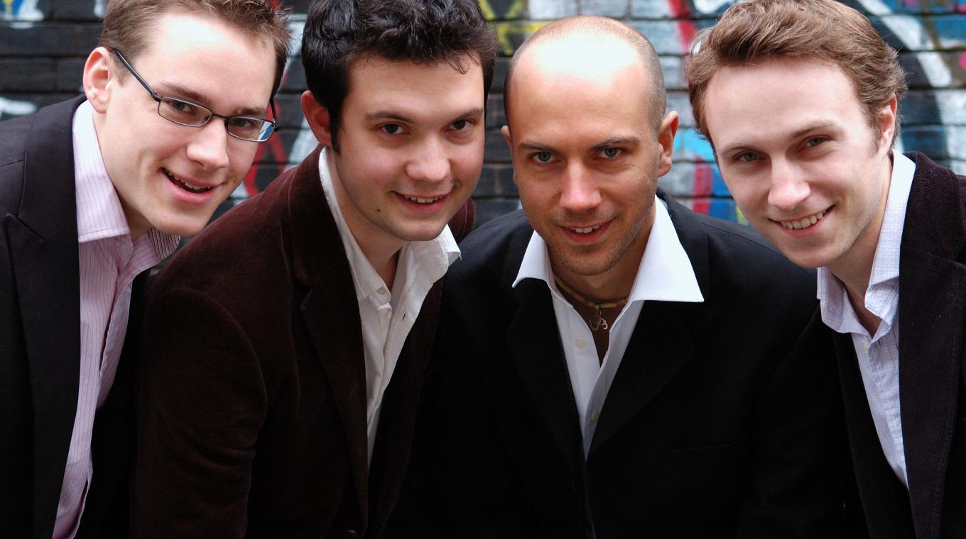 Four men with open shirts and jackets leaning forward together to the camera