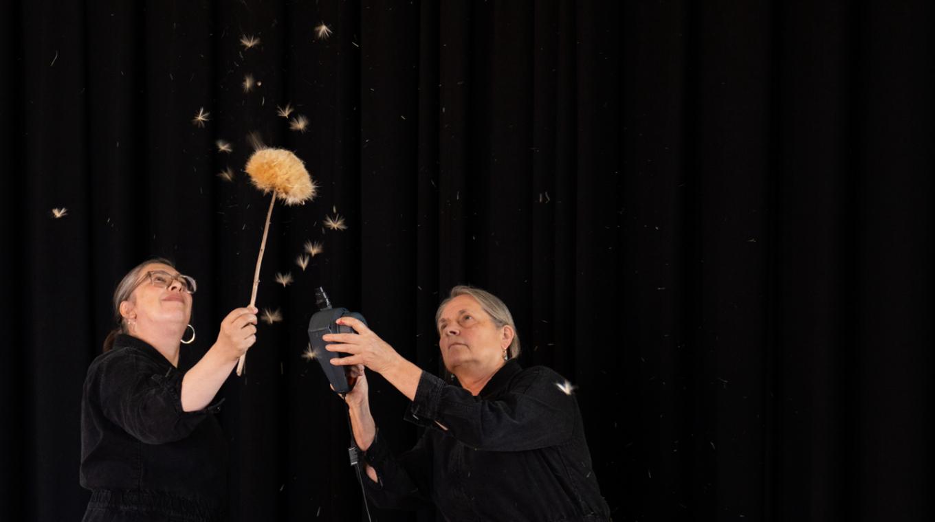 Two women, one holding a plant like a dandelion, and the other a fan which is blowing the seeds from the plant in the air
