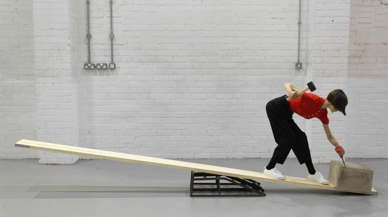 A woman standing on a plank, chpping at a block of concrete