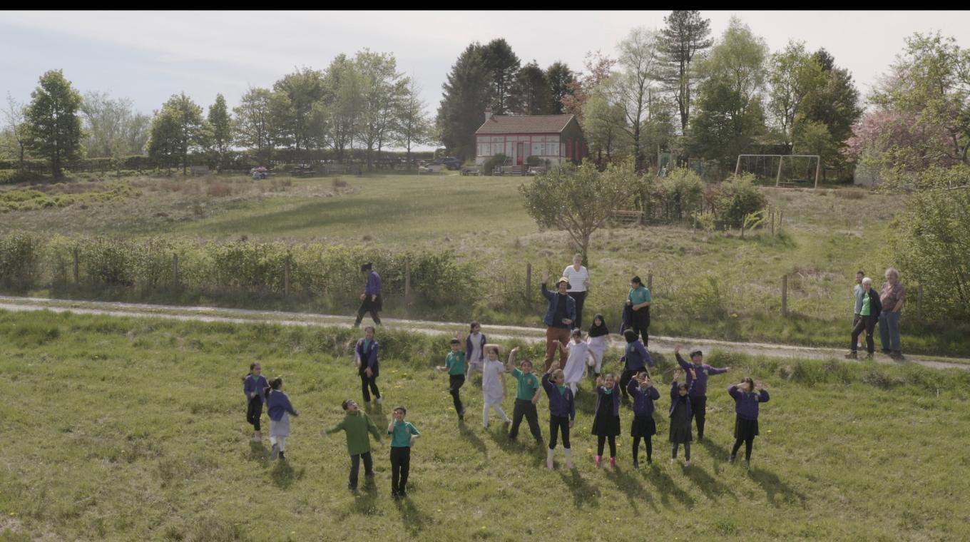 An aerial view of a large group of children waving in a field with a house behind