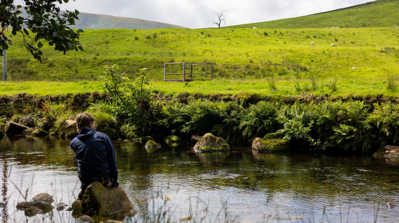 A man sitting on a rock in a river looking out at green fields