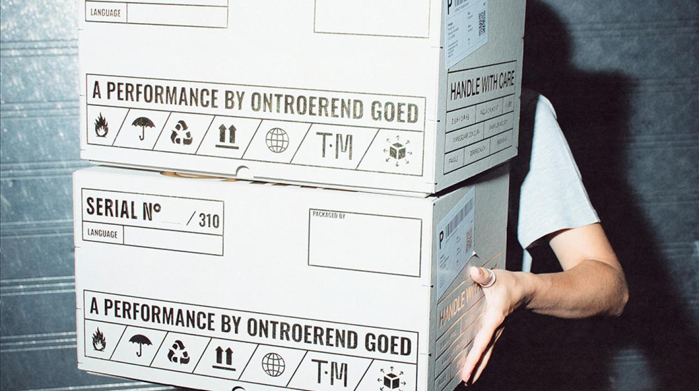 A person hidden behind two white boxes they are carrying with the words "a performance by Ontroerend Goed" on them