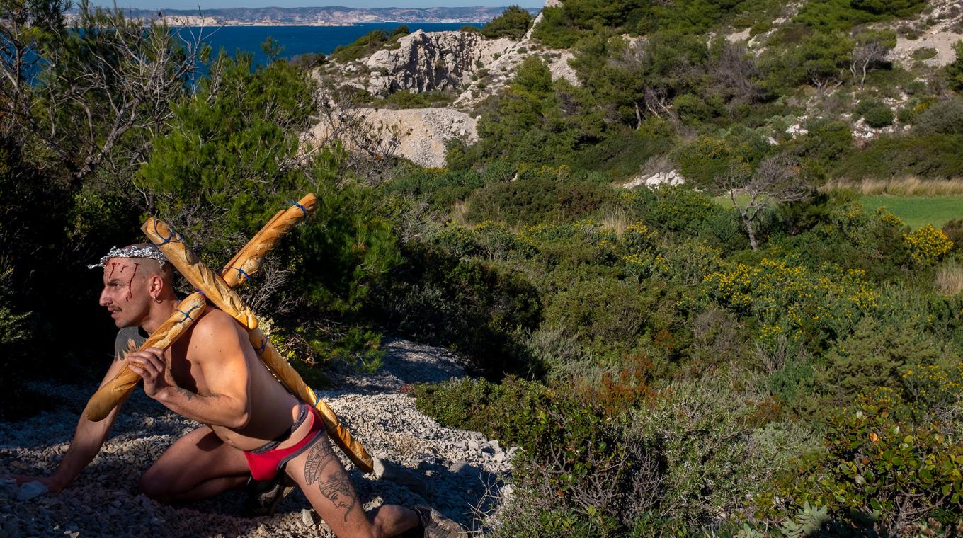 a semi-naked man carrying a cross made of baguettes up a mountain