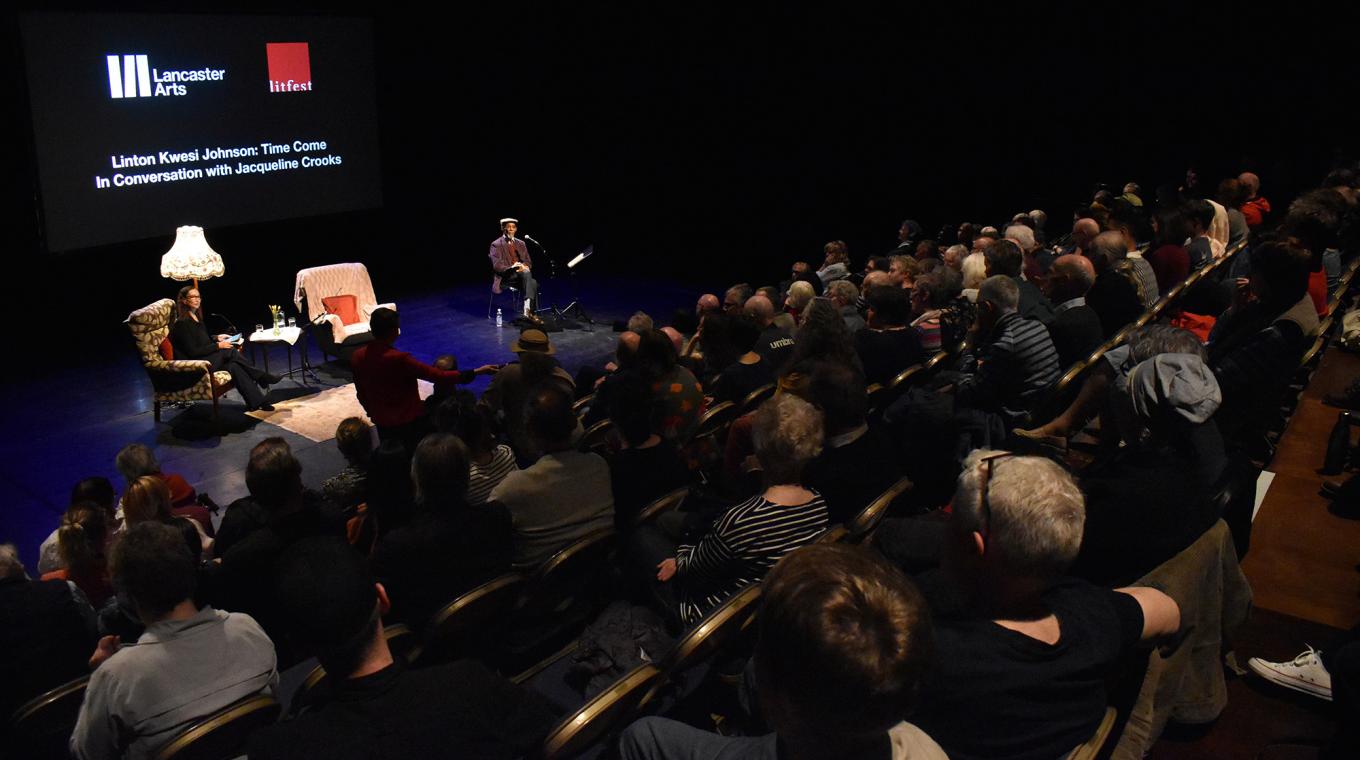 An audience in raked sitting watchiong two people in arms chairs on stage in a theatre