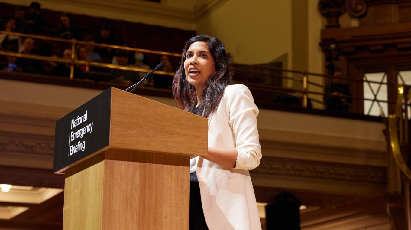 A woman in a white jacket speaking from a podium that has a sign saying "National Emergency Briefing"