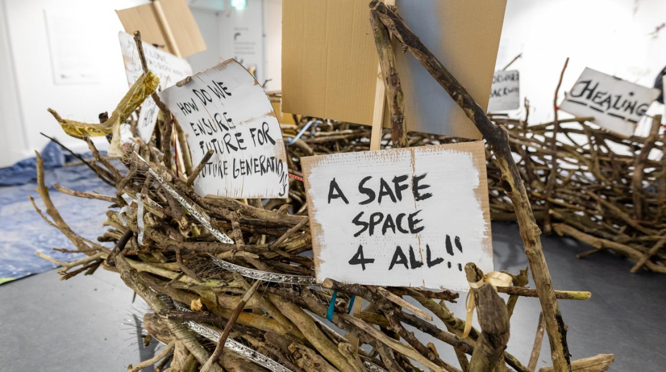A giant nest in a gallery with signs saying "A safe space 4 all" and "how do we ensure future for future generations"
