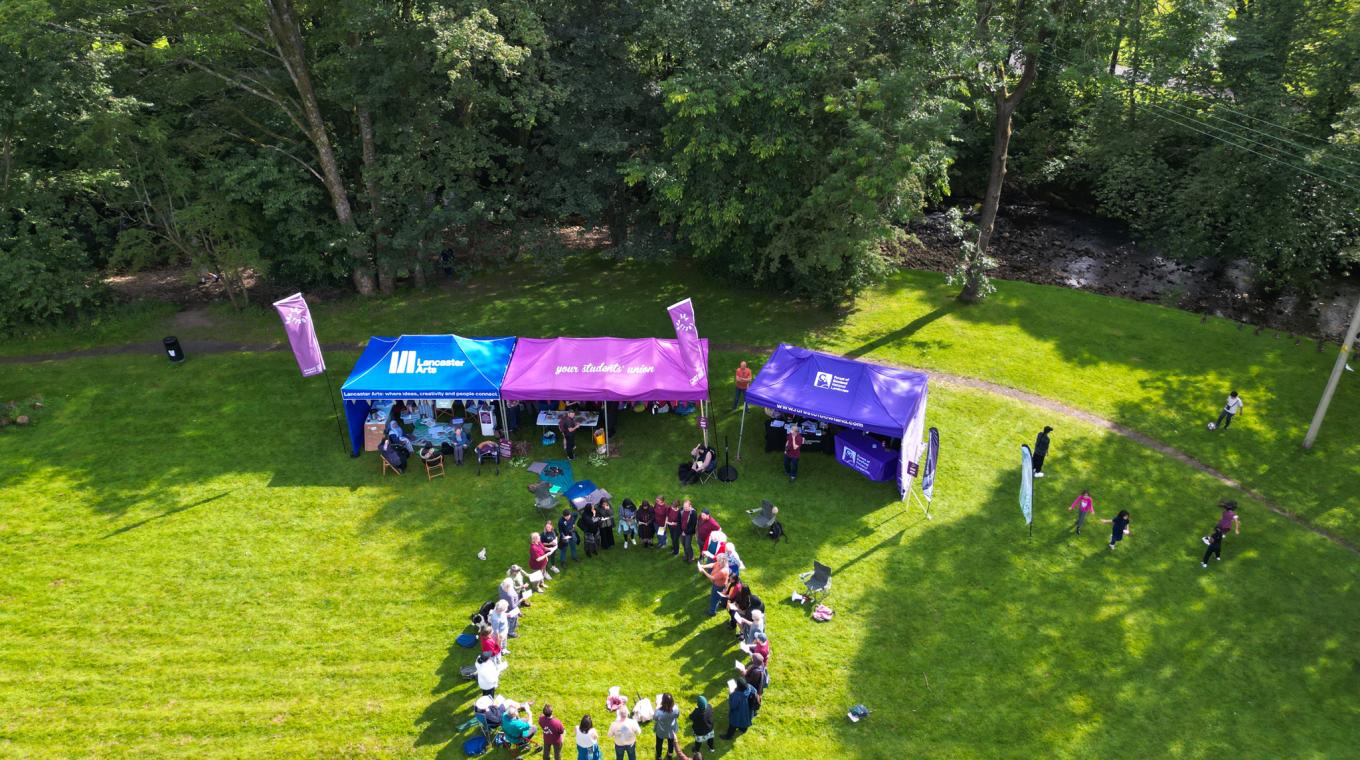 Aerial image of around 30 people standing in a circle on a village green near some some branded gazebos