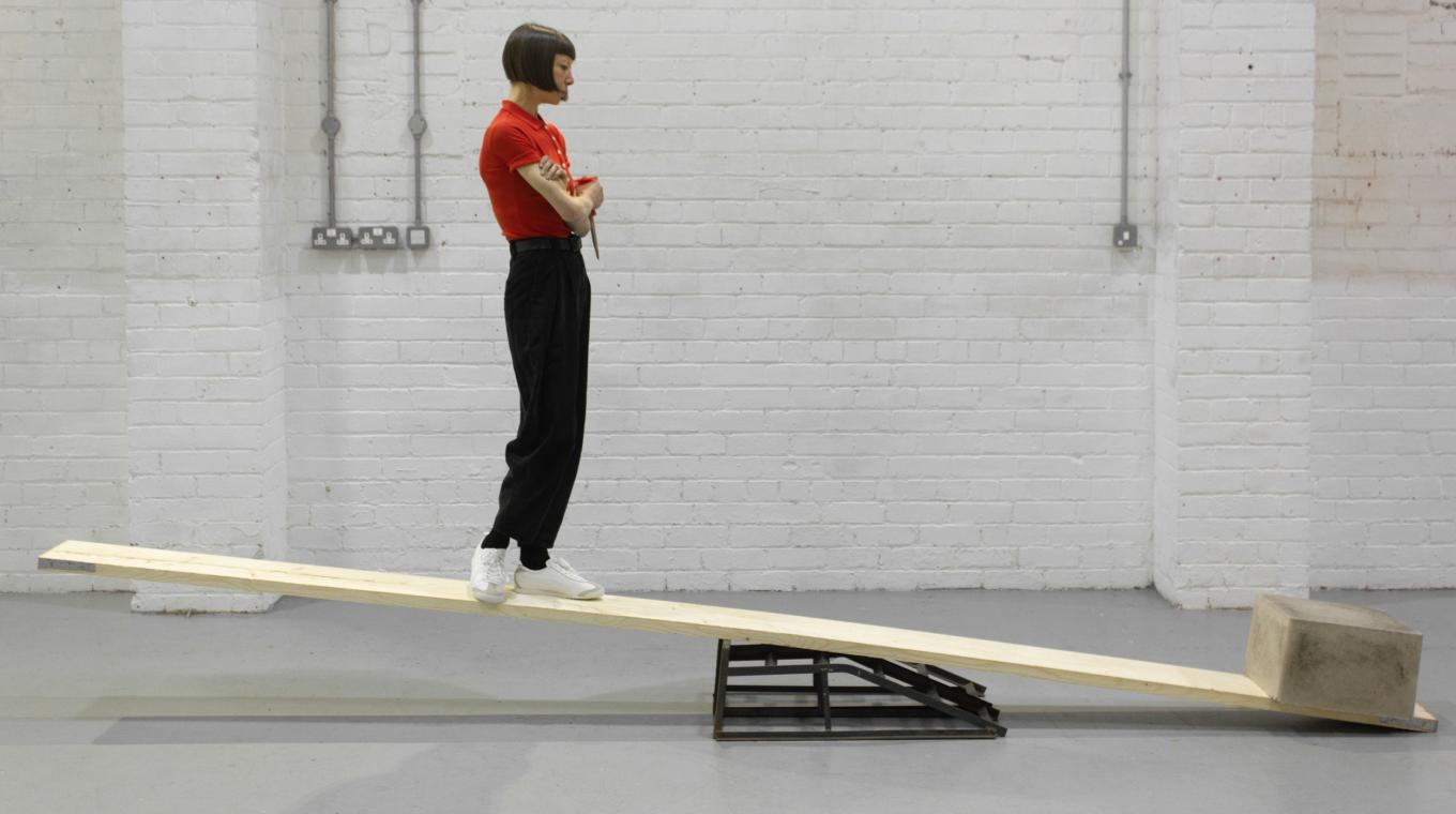 A woman with a red top standing on one end of a plank trying to balance with a concrete block on the other end