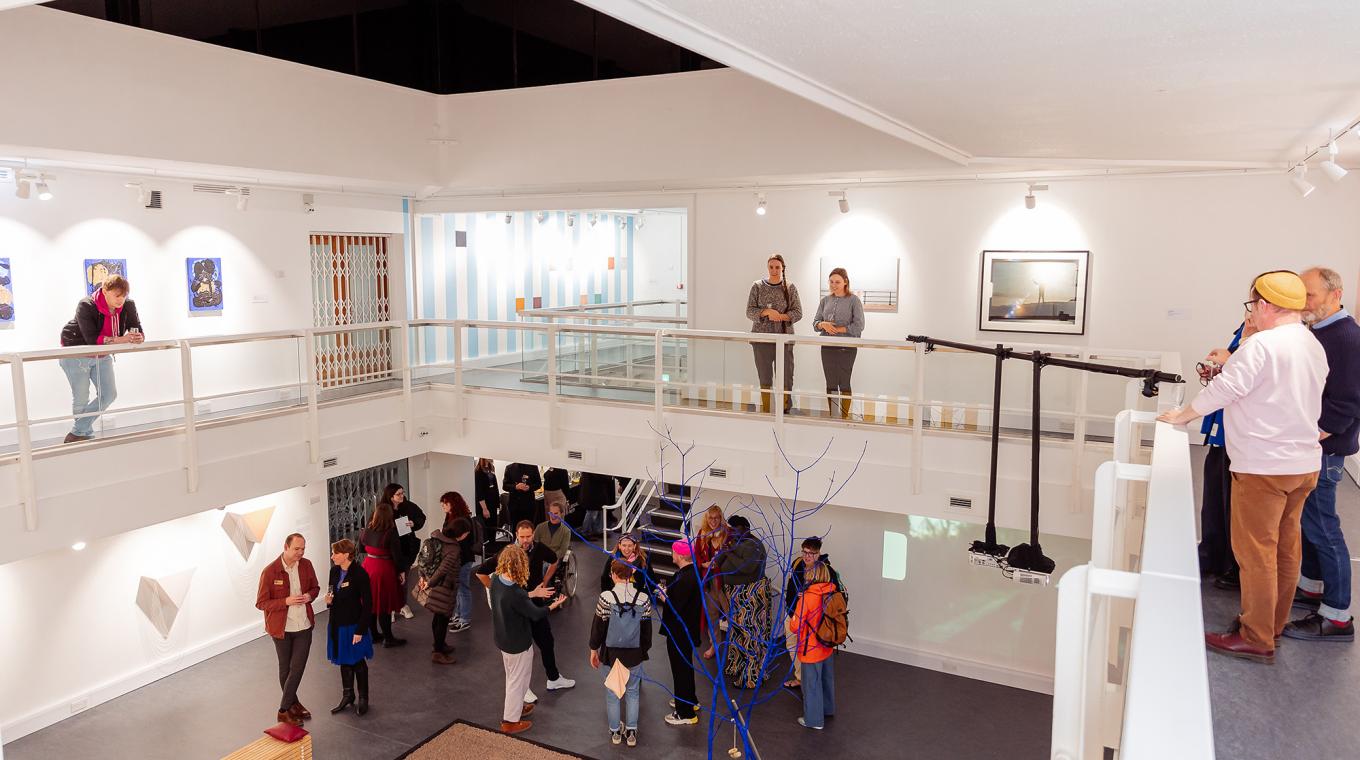 People on two floors in a gallery full of artworks including a blue tree on sand in the centre of the gallery