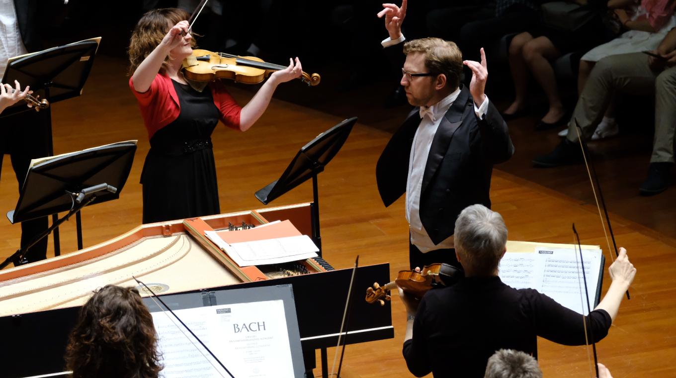 A man conductoing an orchestra with three violinists visible